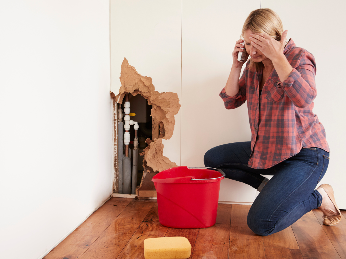 Upset woman on the phone kneeling beside a wall that has been damaged by a plumbing disaster